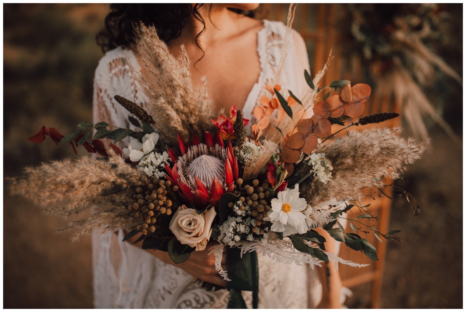 Bride wearing a lace boho dress and holding a bouquet of roses, pampas grass and protea in the desert