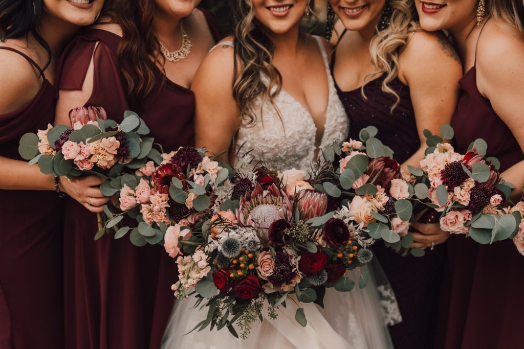 bride and bridesmaids holding burgundy, blush, and peach bouquets with roses, ranunculus and protea