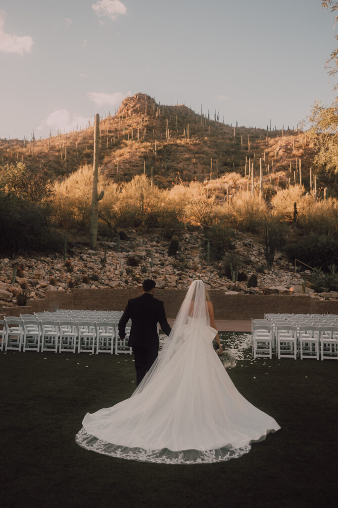 arizona wedding, ceremony space, wedding ideas, veil and wedding dress train, bride and groom walking away from the camera towards the ceremony space surrounded by cactus and desert 